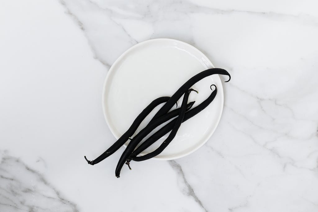 Top view of aromatic vanilla beans on a white plate placed on a marble surface, showcasing minimalist still life photography.